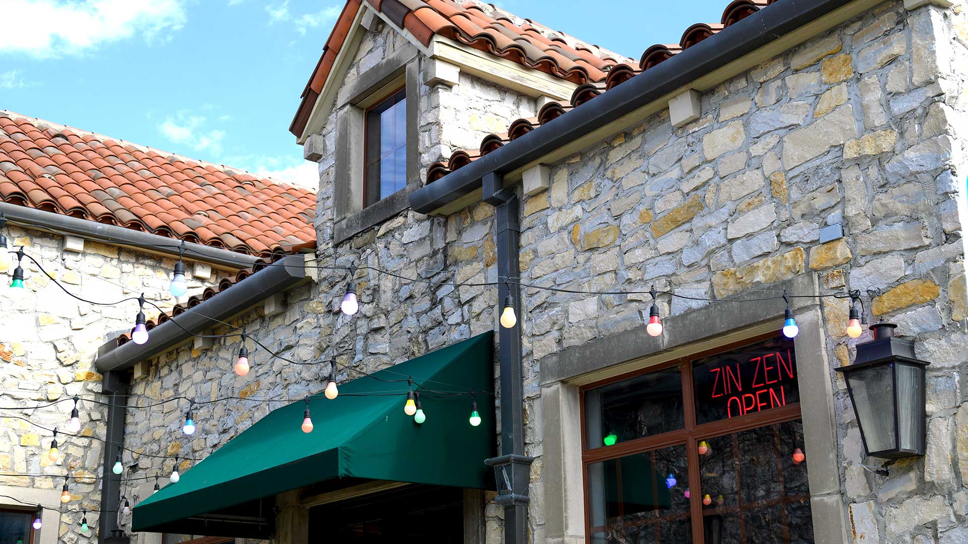 A close up look at the exterior of a restaurant at Adriatica. The building is stone with a green awning over the door. Several lights hang from strings across the walkway.